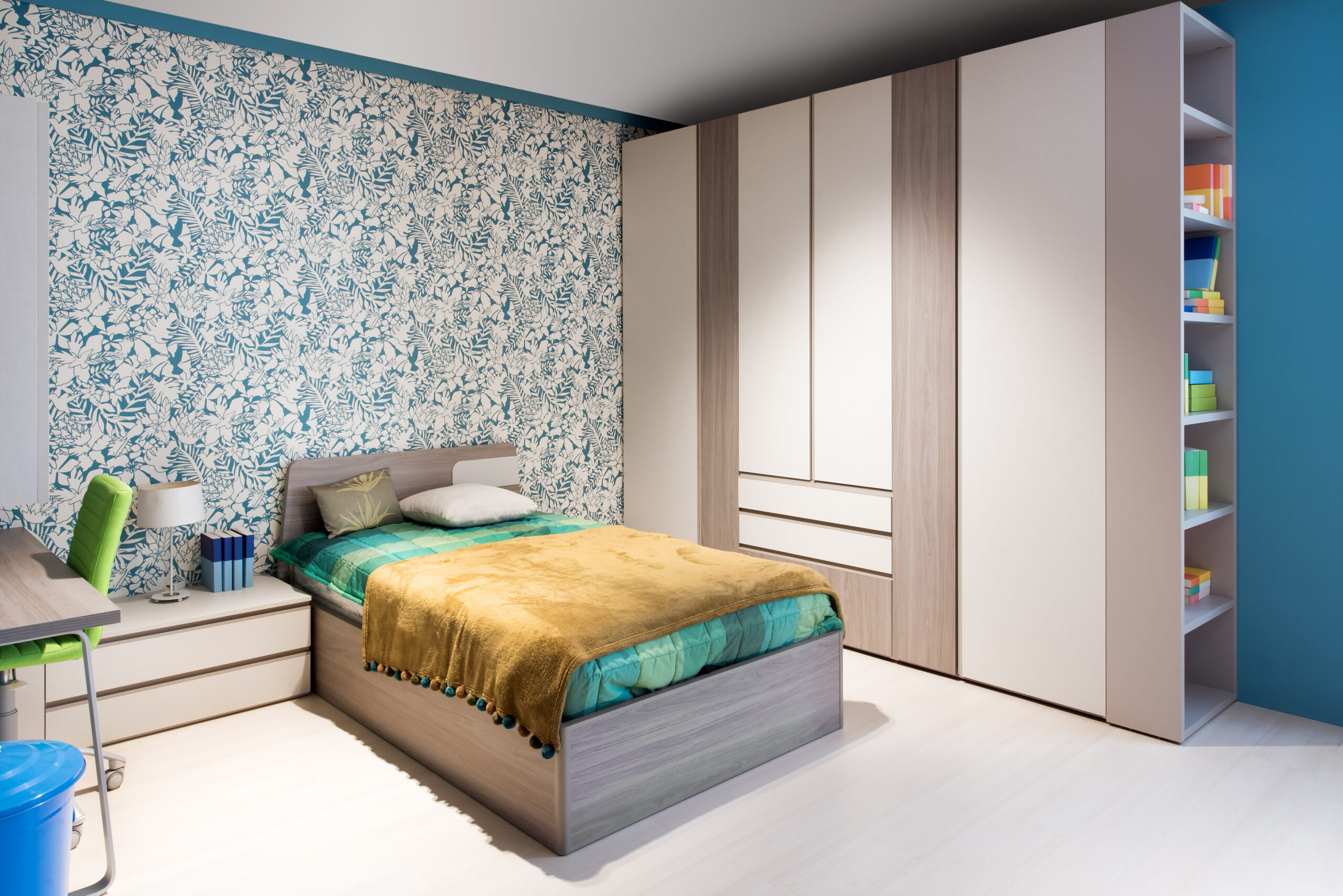 Blue and green themed bedroom interior with feature wall paper, wooden cabinets and a single divan bed alongside a small desk